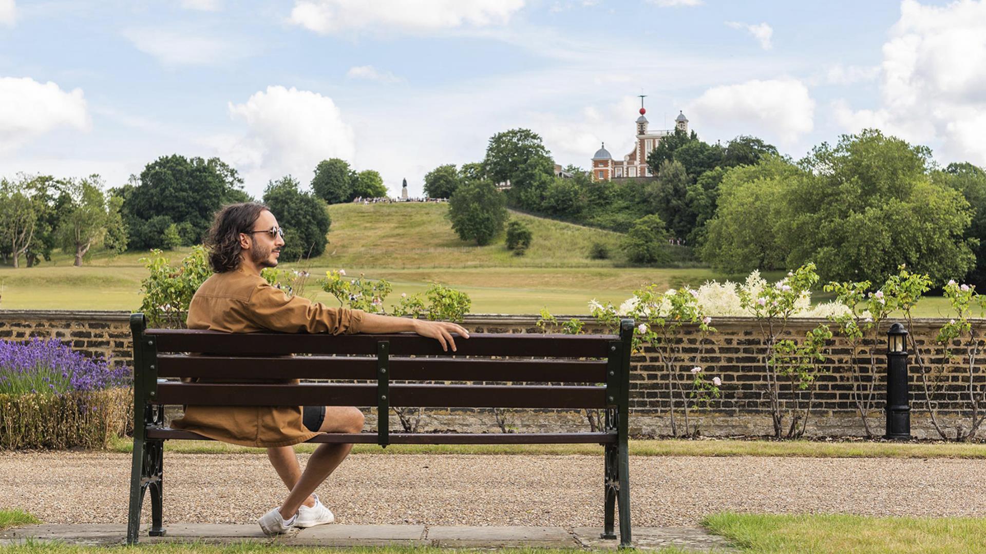 A man sits on a bench in Greenwich Park in front of the Royal Observatory.