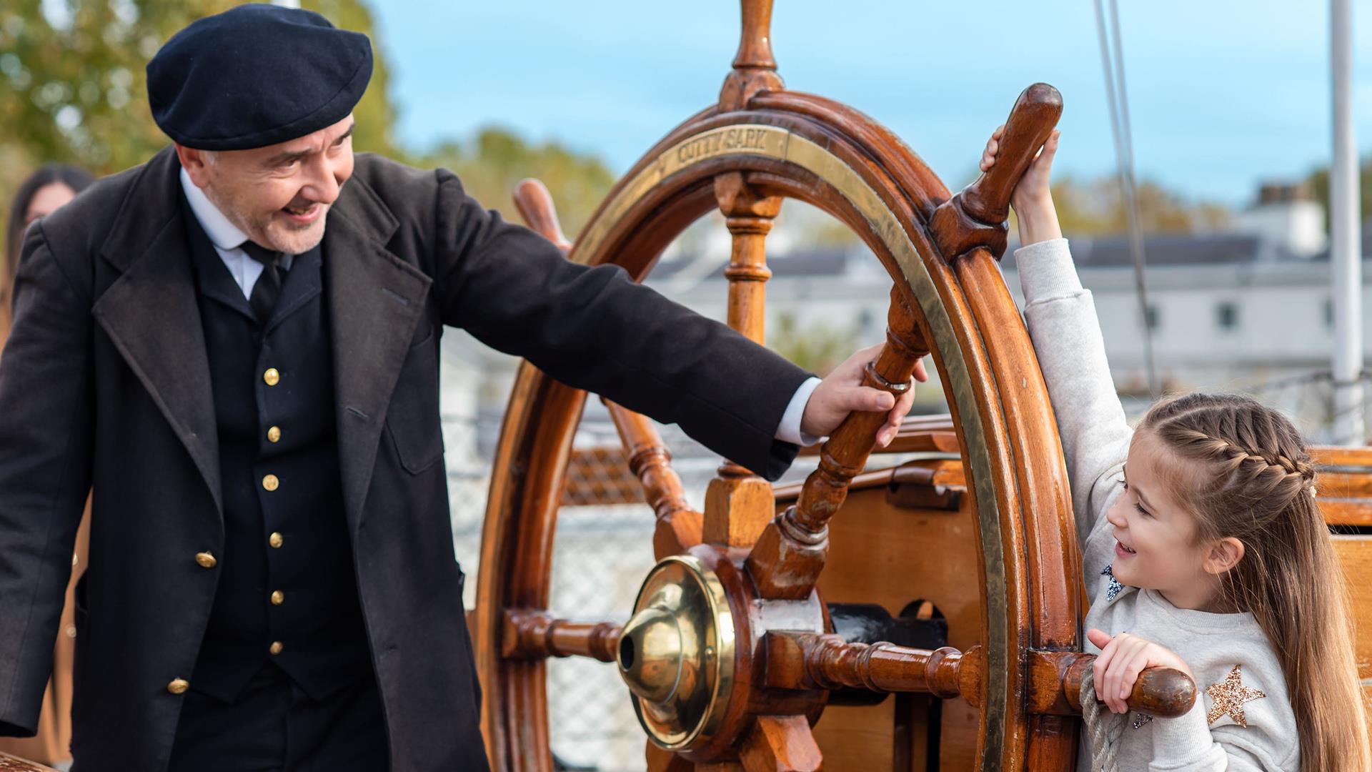 A girl holds on to the wheel of the Cutty Sark in Greenwich while an actor dressed as the captain looks on.