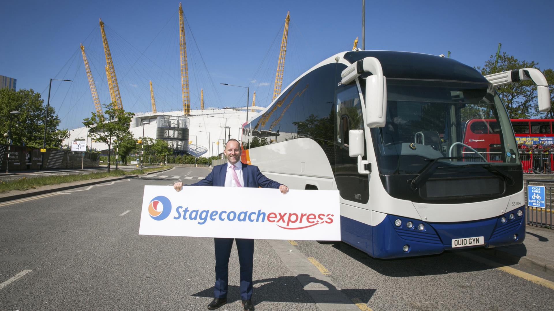 A man holds a sign for Stagecoach in front of a coach with The O2 in the background on Greenwich Peninsula.