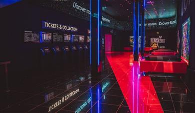 A well taken photo of inside of Cineworld O2, in the picure you can see the tickets and collection area, red and black sofas, movie posters and the en