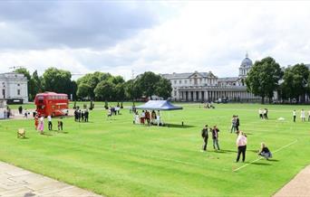 Red bus and people stood on green grass on the Royal Museums Greenwich Grounds