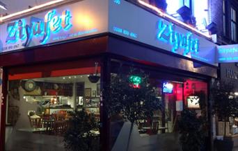 Night view of entrance of Ziyafet Restaurant exterior with big glass window around and name glowing with blue light in the background.