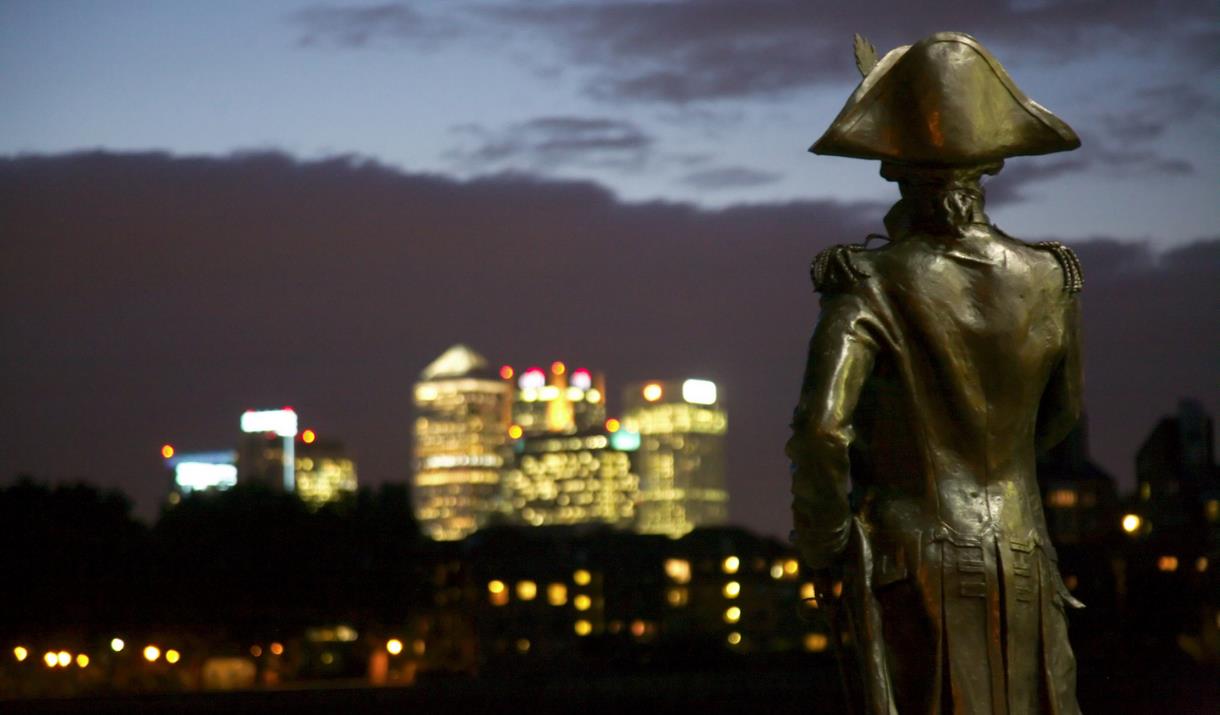 Statue of Lord Nelson overlooks the glittering towers of Canary Wharf at night.