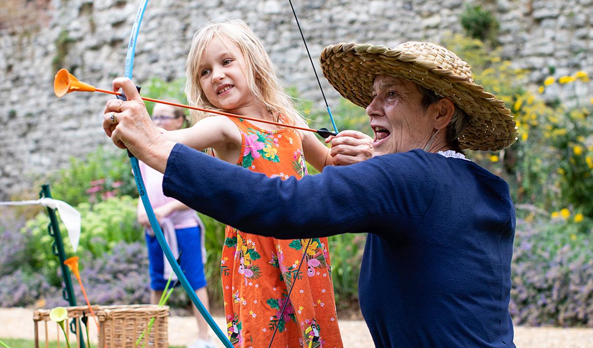 Join Eltham Palace every day of the summer holidays to learn what life was like in medieval times - woman in medieval costume helping a girl with a bo