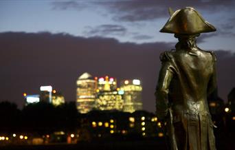 Statue of Lord Nelson overlooks the glittering towers of Canary Wharf at night.