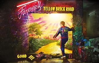 Elton John wearing blue sportswear with pink stripes, standing by the brick wall in the artwork for the Farewell Yellow Brick Road Tour.