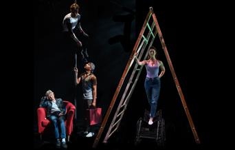 Four performers on a dark stage. The left half of the image is filled with a large ladder, under which a white woman in her thirties stands, balancing