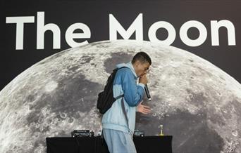 Youth artist with a mic in his hand facing right side in front of 'The Moon' banner.